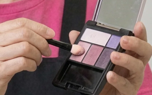 A hand loading a brush with pink eyeshadow from a palette.